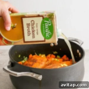 A person pouring a carton of chicken broth into a large pot containing chicken and vegetables.