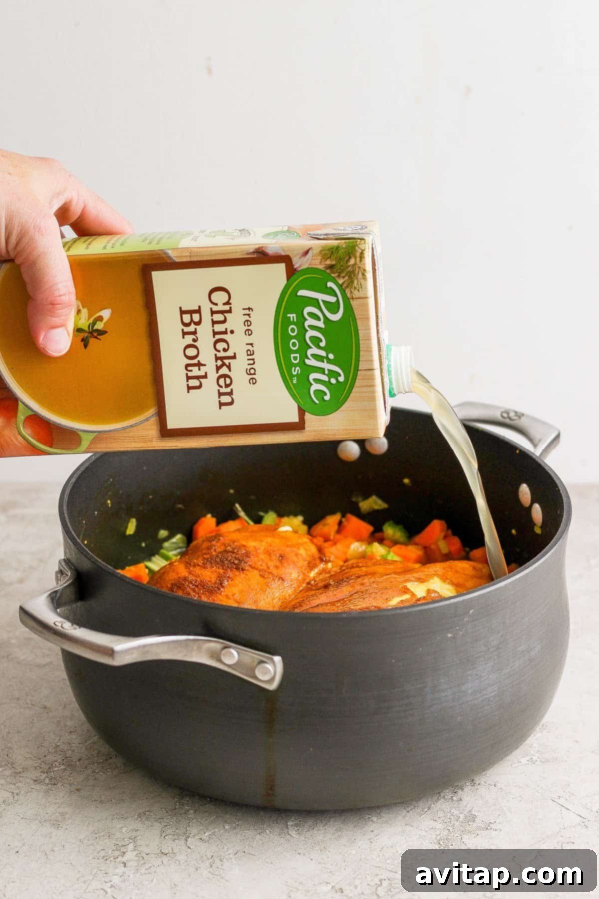 A hand pouring chicken broth from a carton into a pot of sautéed vegetables and chicken.