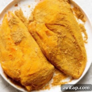 Two pieces of chicken seasoned with golden spices on a plate, ready for cooking.