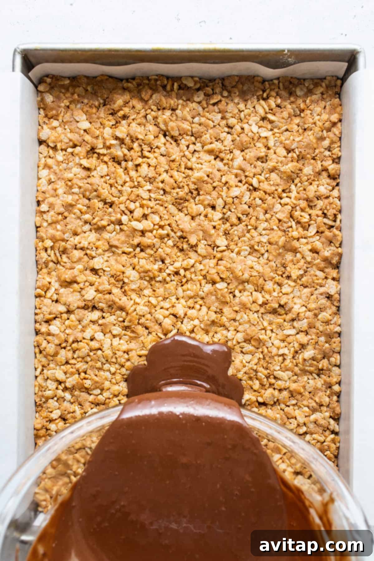 Protein Power Scotcheroos 6 A luscious chocolate sauce being carefully poured over a base of crisp rice cereal in a baking dish, creating the iconic two-layer scotcheroo.