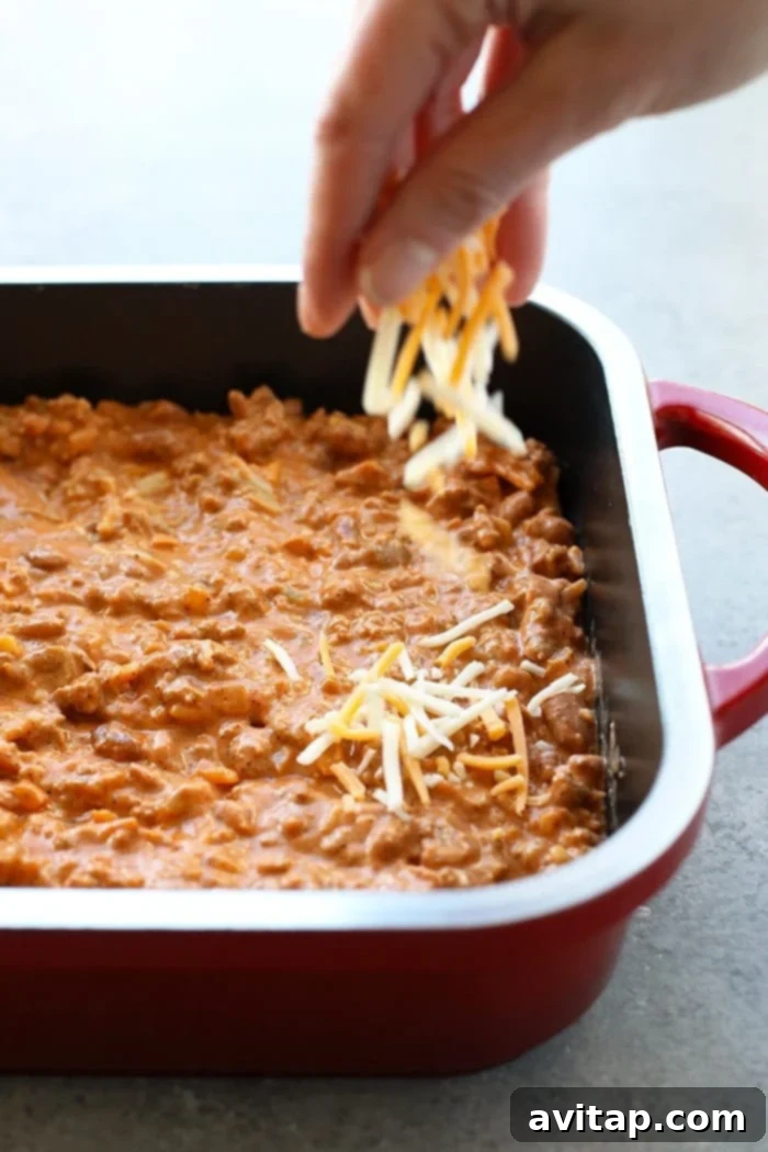 Make this winning chili cheese dip for the big game and your guests will gobble it up. It really is the best chili cheese dip recipe! A hand sprinkling generous amounts of shredded cheese over a casserole dish filled with a savory bean dip, ready for baking.