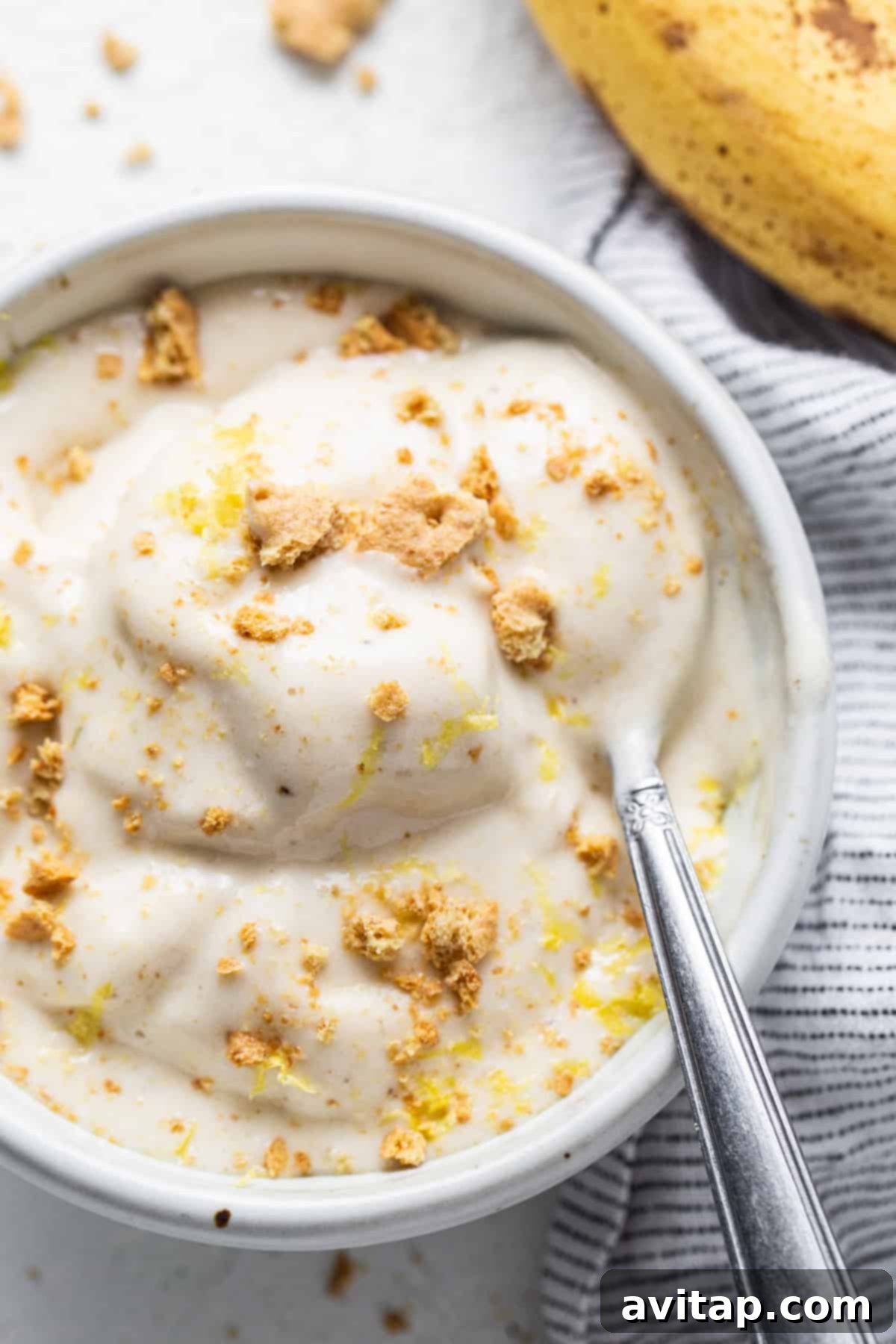 Zesty Lemon Cheesecake Nice Cream 2 A bowl of creamy lemon cheesecake nice cream, topped with graham cracker crumbs and fresh lemon zest, with a spoon resting in it.