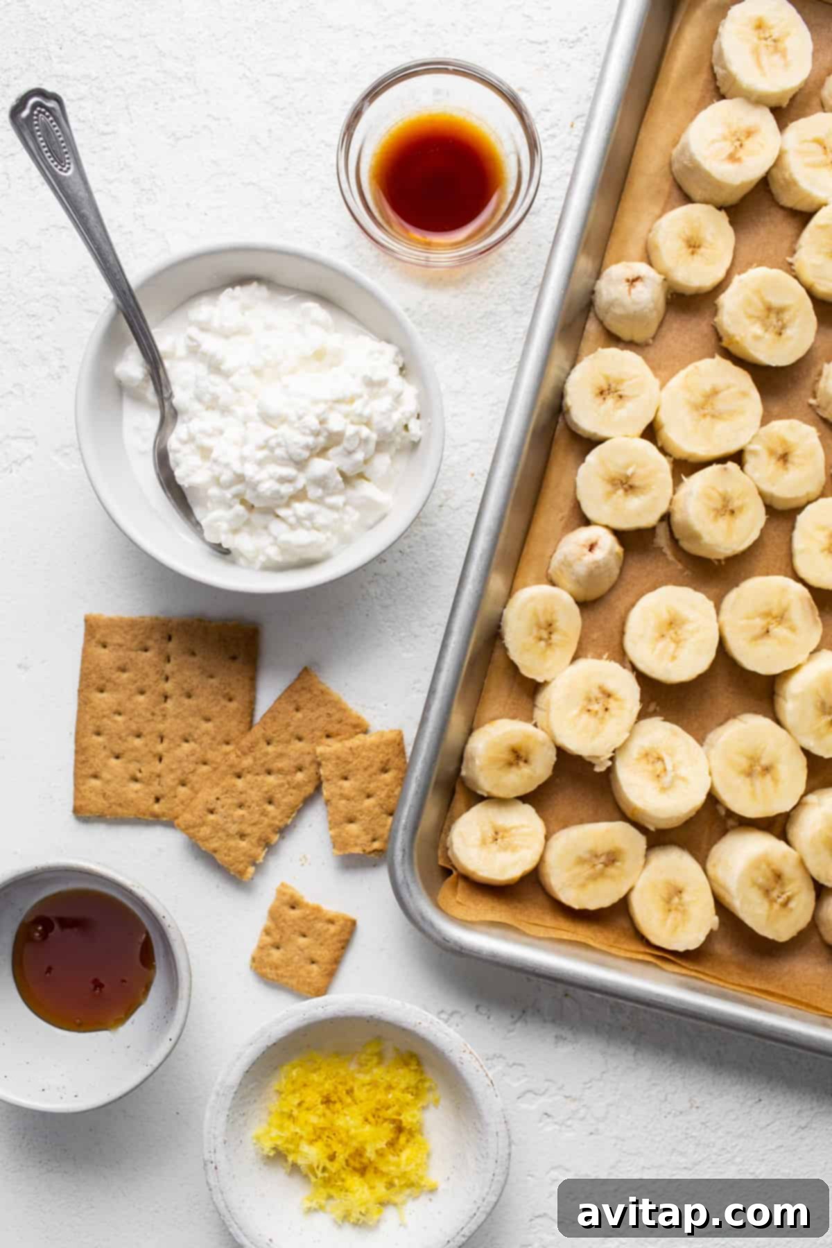 Zesty Lemon Cheesecake Nice Cream 3 A collection of ingredients for lemon cheesecake nice cream including bananas, a block of cottage cheese, a lemon, and graham crackers on a tray, with a spoon.