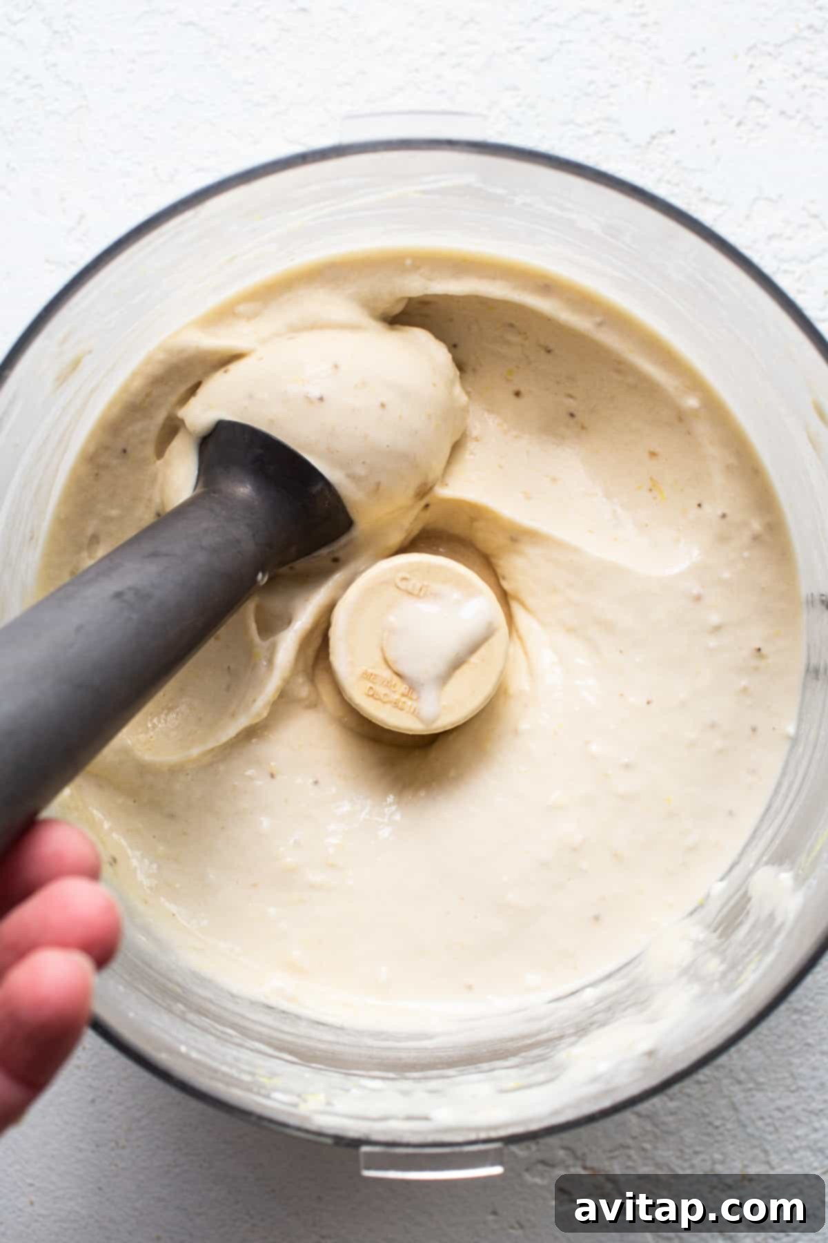 Zesty Lemon Cheesecake Nice Cream 5 A hand holding a food processor tamper, pushing down a white mixture inside the processor.