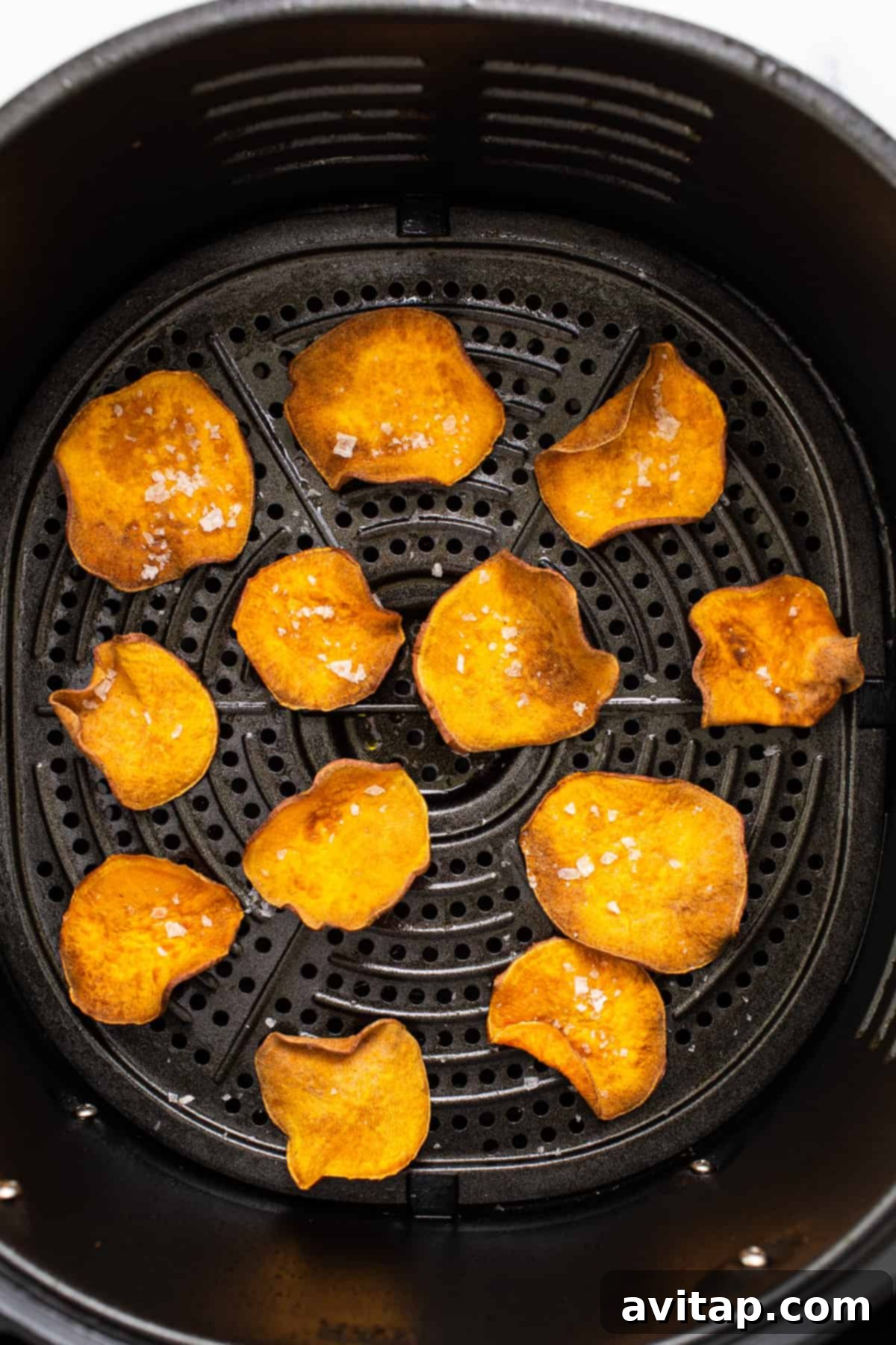 A close-up view of various thin slices of sweet potato being cooked to perfection inside an air fryer basket.