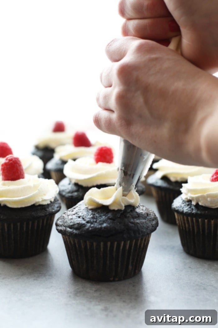 Healthier chocolate cupcakes being piped with creamy vanilla frosting and topped with a fresh raspberry, showing the decorating process.