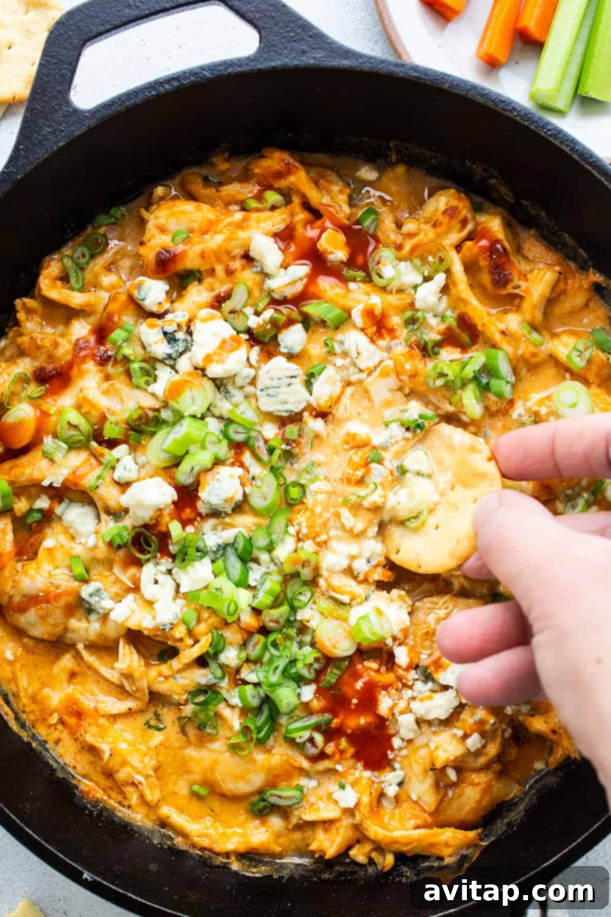 A person dipping a cracker into a hot skillet of whipped cottage cheese buffalo chicken dip, garnished with fresh green onions and blue cheese.