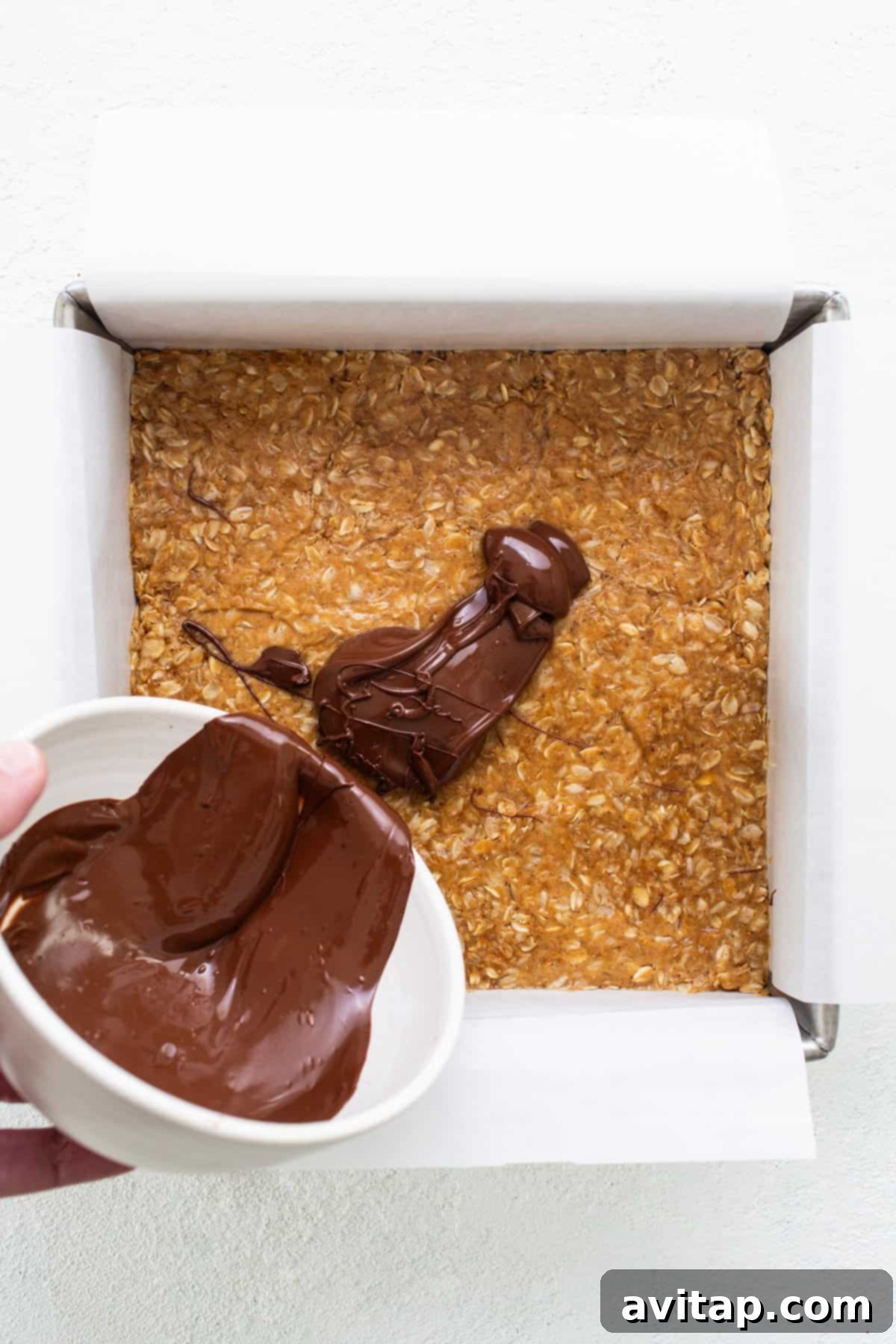 Ultimate Peanut Butter Cup Oatmeal Bars 4 Hands pouring melted chocolate from a bowl over a pressed oat mixture in a baking pan, creating the top layer.
