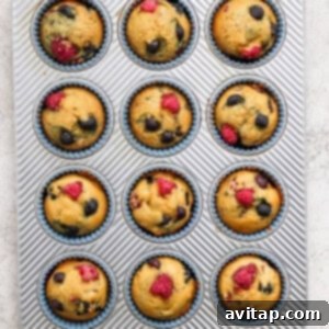 Freshly baked Greek Yogurt Berry Muffins cooling in a muffin tin, garnished with scattered raspberries and blueberries.