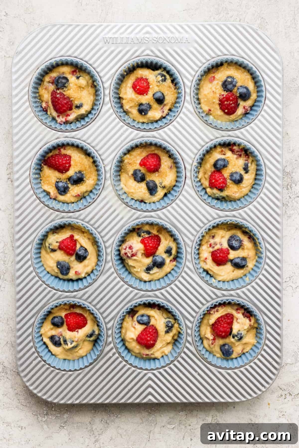A muffin tin generously filled with unbaked Greek Yogurt Berry Muffin batter, topped with extra berries, ready for the oven.