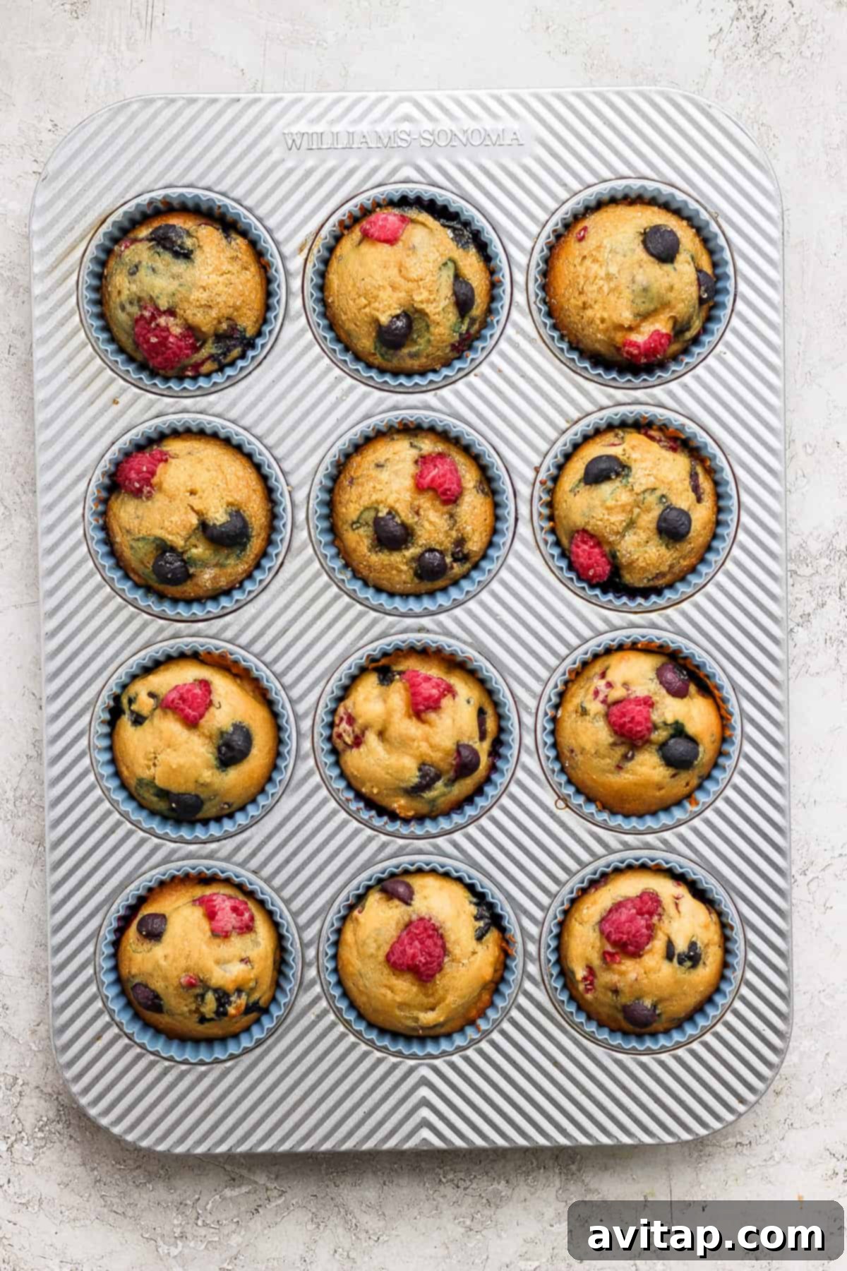 Freshly baked Greek Yogurt Berry Muffins cooling in a muffin tin, topped with scattered raspberries and blueberries.