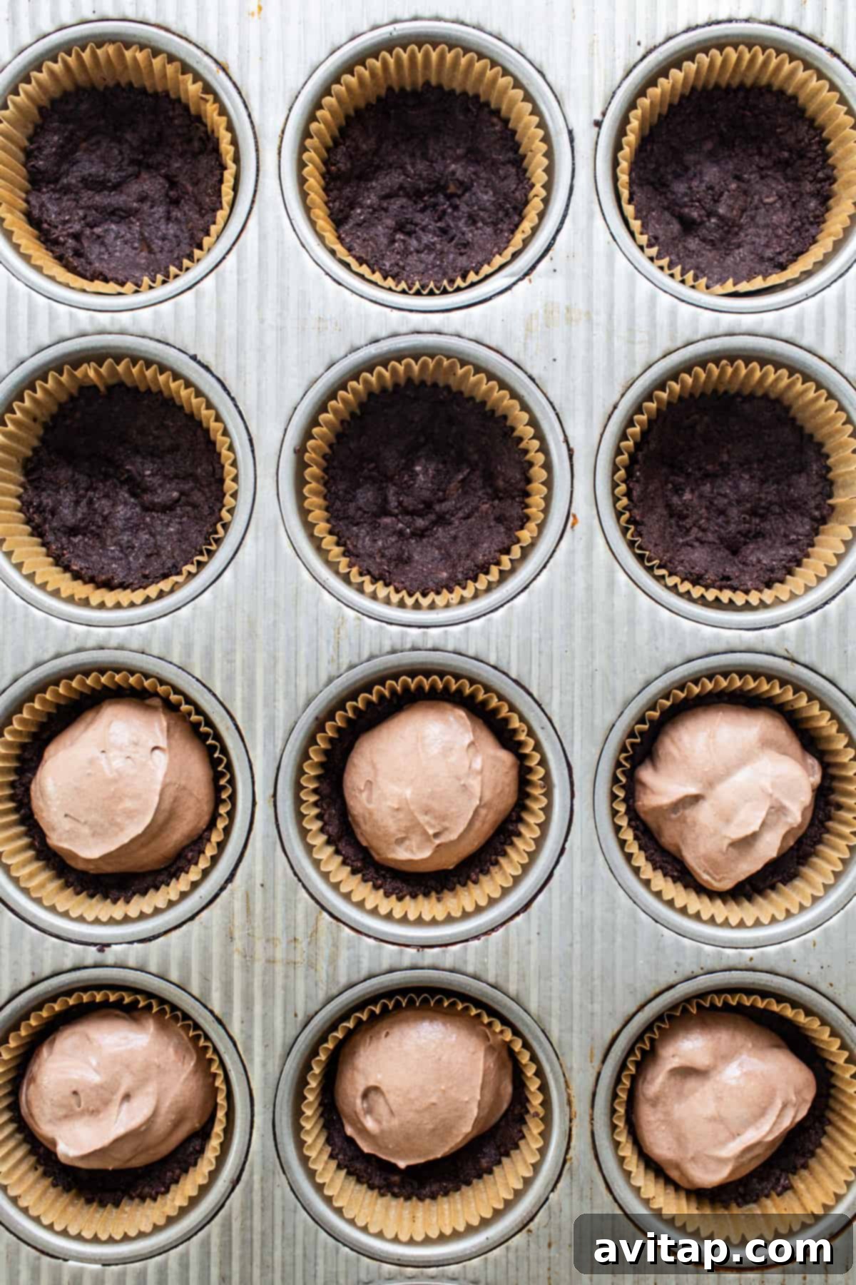 A close-up shot of chocolate cupcake liners neatly arranged within a muffin tin, some already filled with a dark cookie crust.