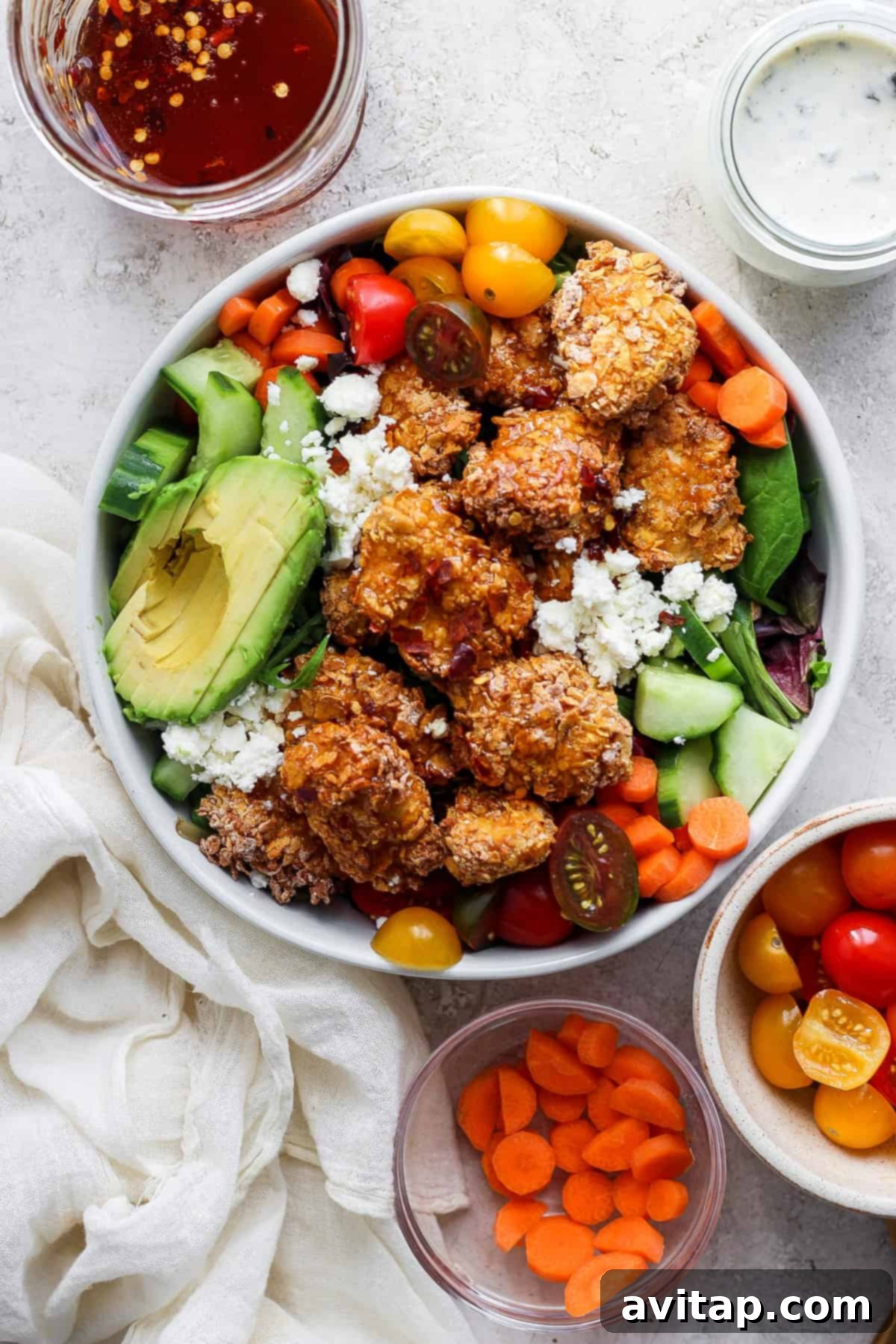 A vibrant salad bowl featuring crispy hot honey chicken, fresh mixed greens, avocado, cherry tomatoes, cucumbers, carrots, and crumbled feta cheese, with two small dressing bowls on the side.
