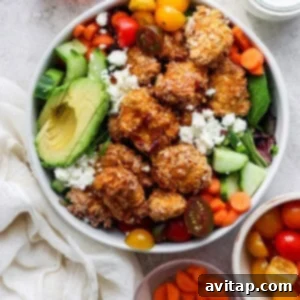 A colorful salad bowl with quinoa, mixed greens, avocado slices, cherry tomatoes, cucumbers, carrots, and topped with crumbled cheese and breaded chicken pieces, accompanied by two small bowls.