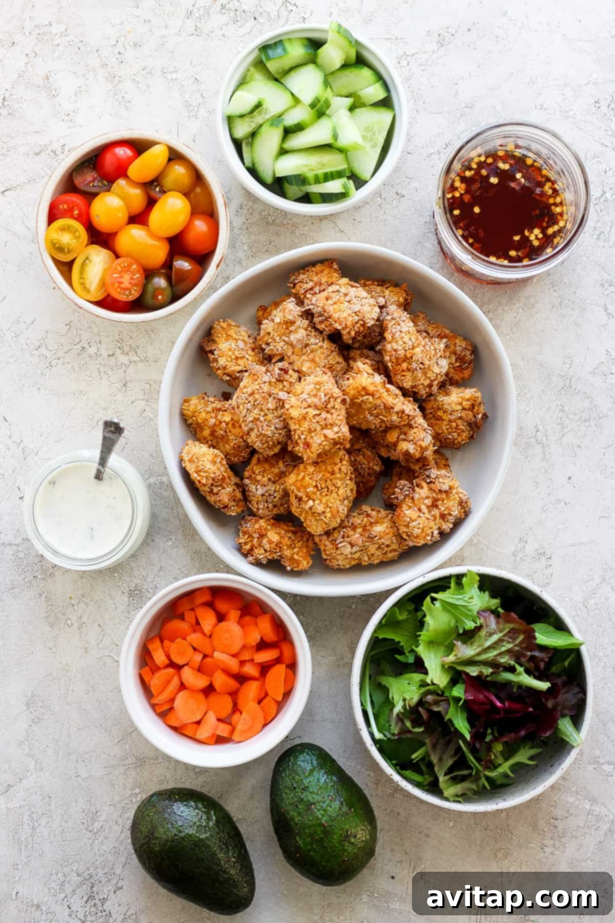 An array of fresh, healthy ingredients laid out, including uncooked breaded chicken pieces, bowls of vibrant vegetables, mixed greens, sliced avocado, and creamy dips, ready for salad assembly.