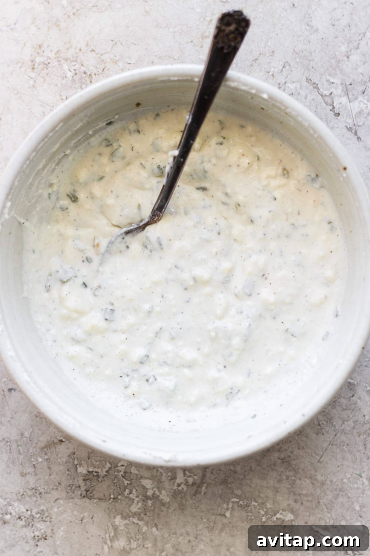 A elegant bowl of creamy herb dressing with a silver spoon resting inside, all placed on a richly textured surface.