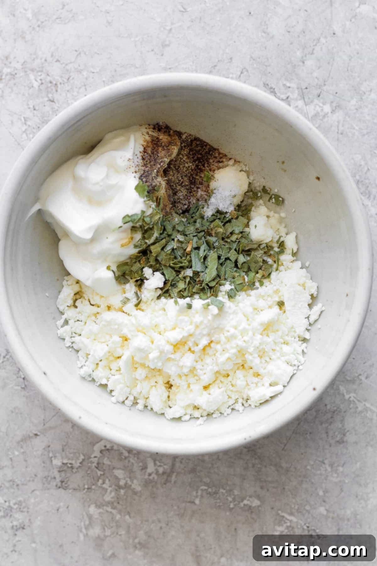 A ceramic bowl containing a creamy mixture of crumbled feta cheese, various seasonings, and Greek yogurt, set against a rustic textured background.