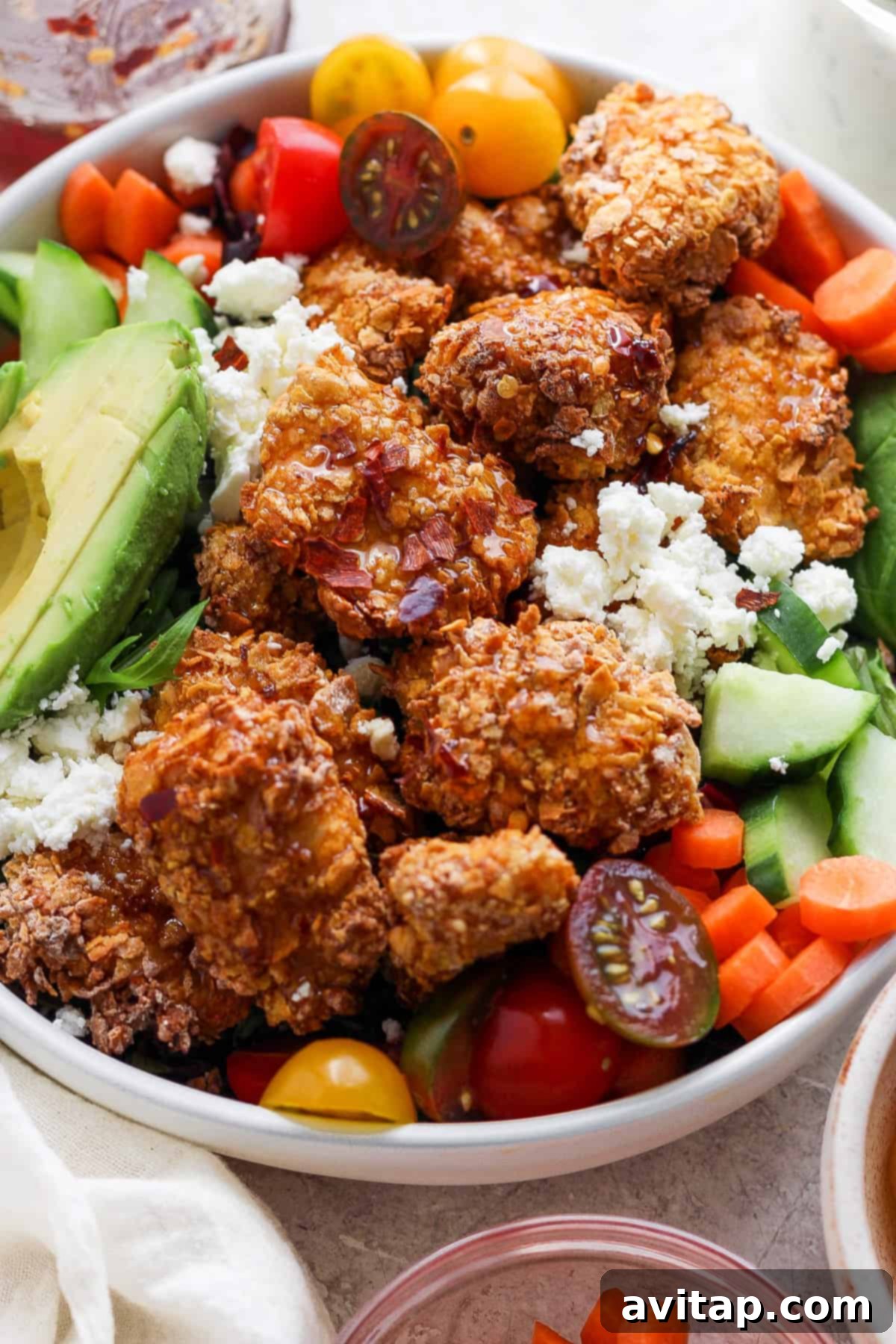 A colorful and appetizing salad bowl brimming with crispy fried chicken pieces, creamy avocado slices, bright cherry tomatoes, cool cucumbers, crisp carrots, and crumbled feta cheese.