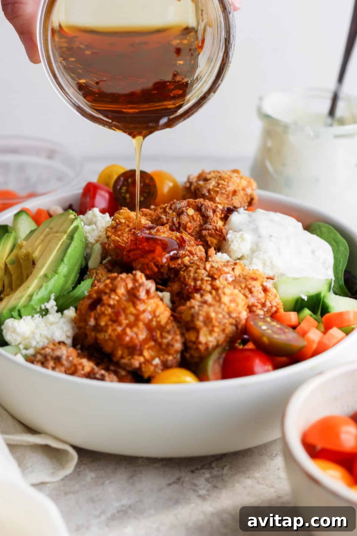 A stream of creamy salad dressing being poured over a visually appealing bowl of salad, featuring crispy chicken, avocado, cherry tomatoes, and mixed greens.