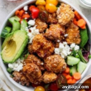A bowl containing fried chicken, avocado slices, cherry tomatoes, sliced cucumbers, carrots, and leafy greens, topped with crumbled cheese.