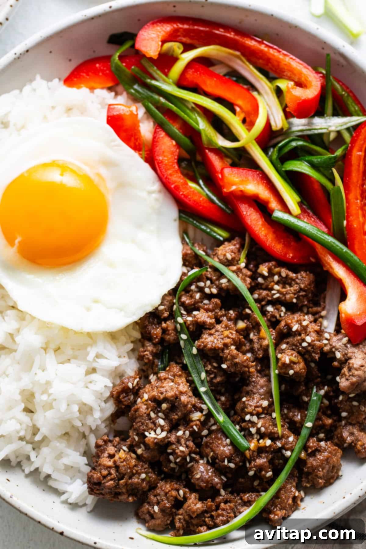 Korean Beef Bowls 2 A vibrant bowl featuring white rice, topped with a perfectly cooked sunny-side-up egg, seasoned Korean ground beef, bright red bell pepper slices, and fresh green onions, all garnished with toasted sesame seeds.