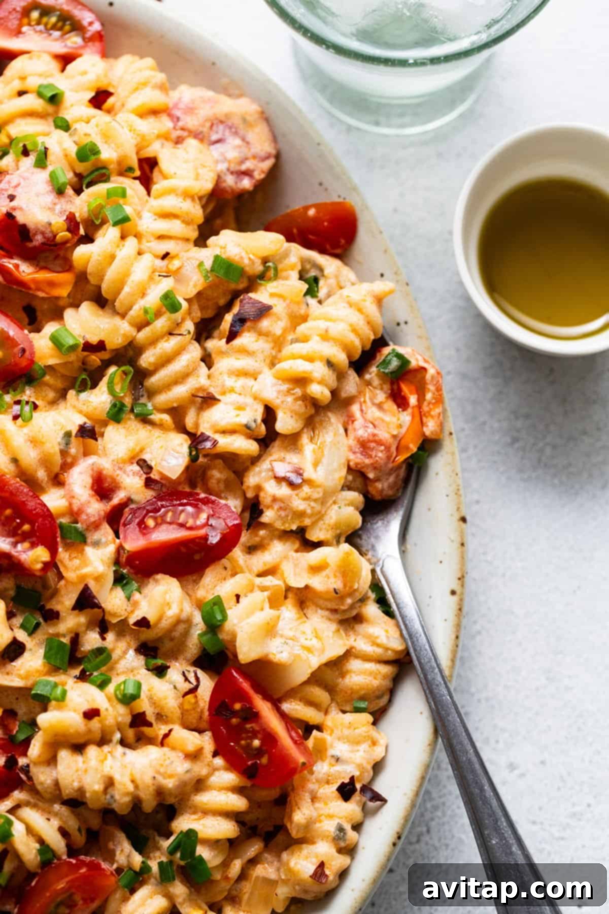 A gourmet bowl of creamy pasta generously topped with sliced sausage, vibrant cherry tomatoes, and fresh chopped green onions, served with a side of golden olive oil.