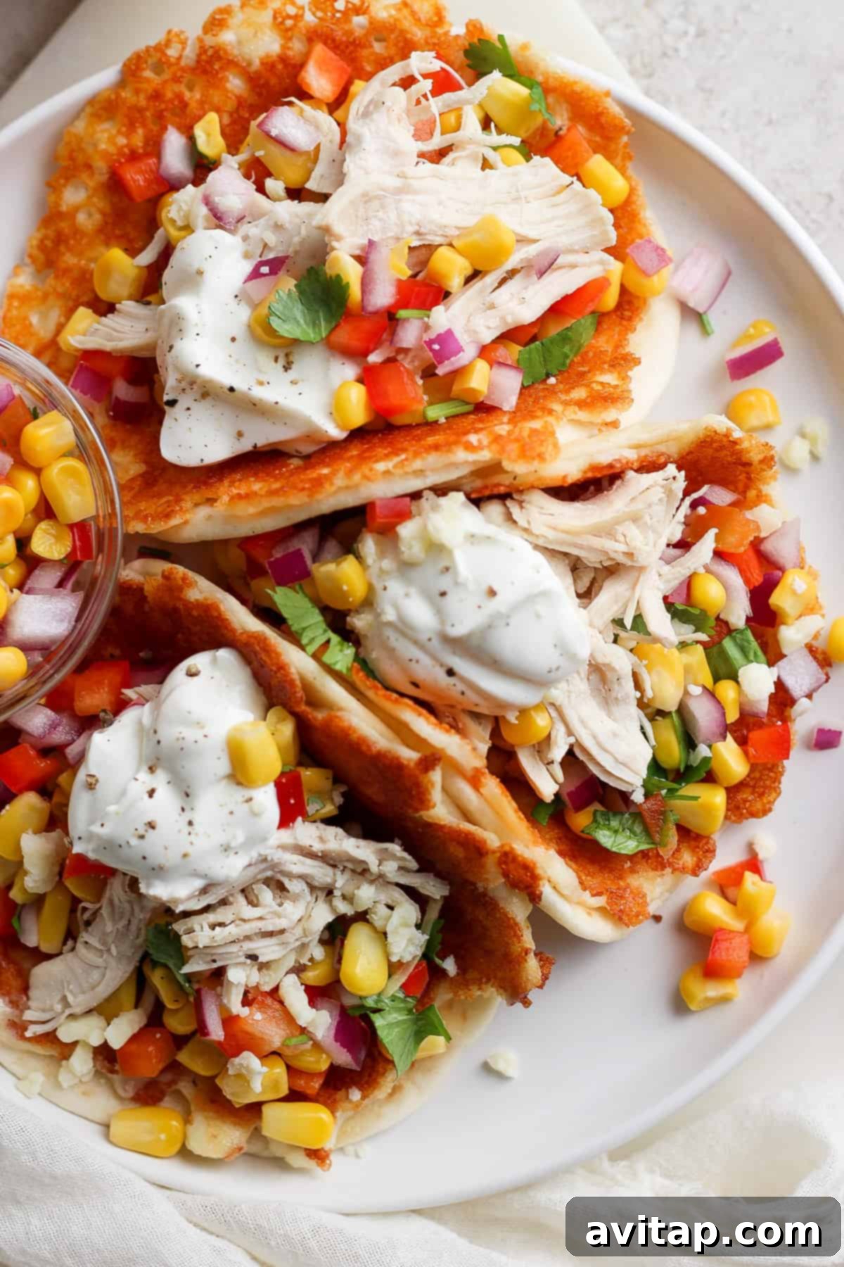 A plate containing chicken tacos garnished with corn, diced tomatoes, red onion, cilantro, and a dollop of sour cream, served on a white surface.