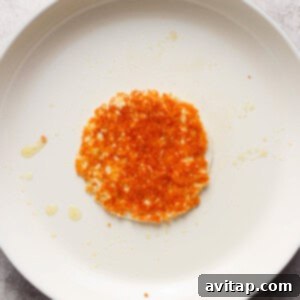A single, fried hash brown resting on a white, circular non-stick pan with a red handle, displayed on a grey surface with a few scattered crumbs and oil droplets.