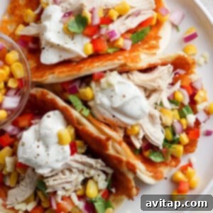 A plate containing chicken tacos garnished with corn, diced tomatoes, red onion, cilantro, and a dollop of sour cream, served on a white surface.