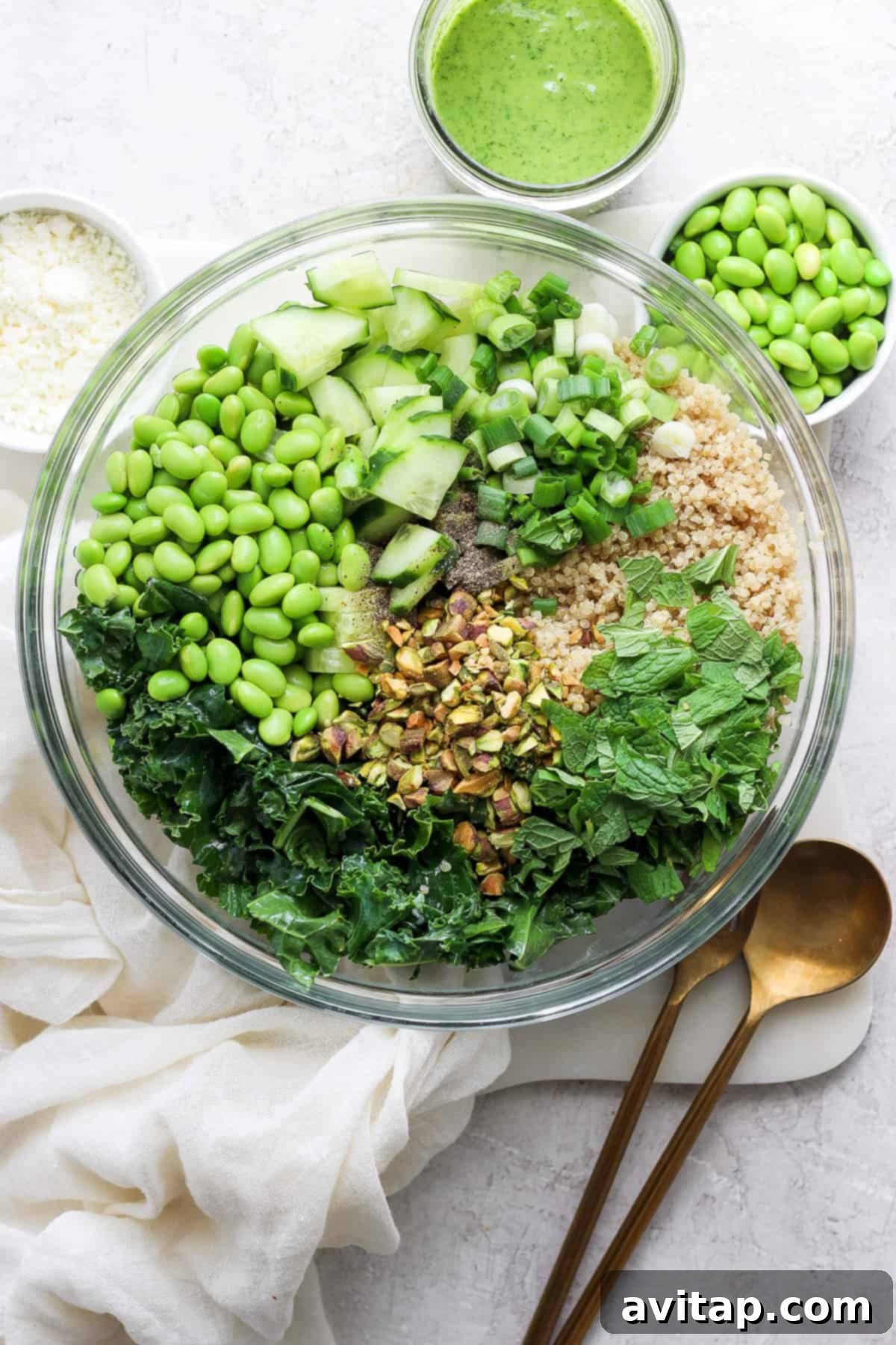 A beautifully arranged bowl filled with a colorful array of green ingredients, including vibrant kale, crisp cucumber slices, bright green peas, and crunchy chopped pistachios. A light dusting of crumbled feta cheese adorns the top, with a jar of creamy green dressing and a serving spoon placed artfully on a white surface, inviting one to indulge in this healthy Green Goddess Quinoa Salad.