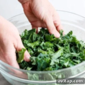 Hands gently massaging chopped kale with olive oil in a glass bowl to tenderize the leaves and enhance their flavor.