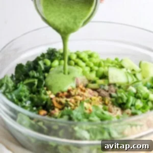 Creamy green goddess dressing being generously poured over a vibrant bowl of quinoa salad with kale, cucumbers, and green peas, ready to be thoroughly tossed.