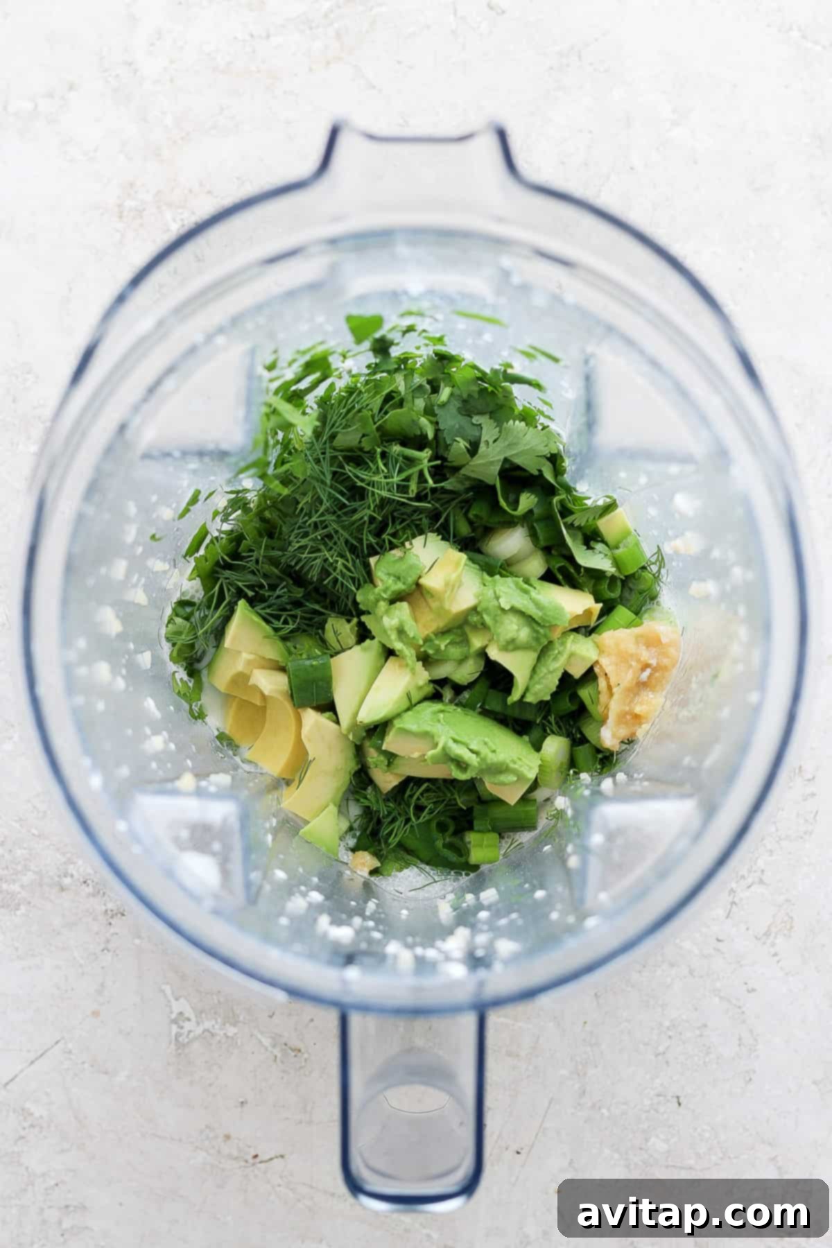 A close-up shot of a blender jar filled with a vibrant mix of fresh ingredients like bright green herbs, creamy avocado, and various liquid components, poised for blending into a delicious and healthy dressing.