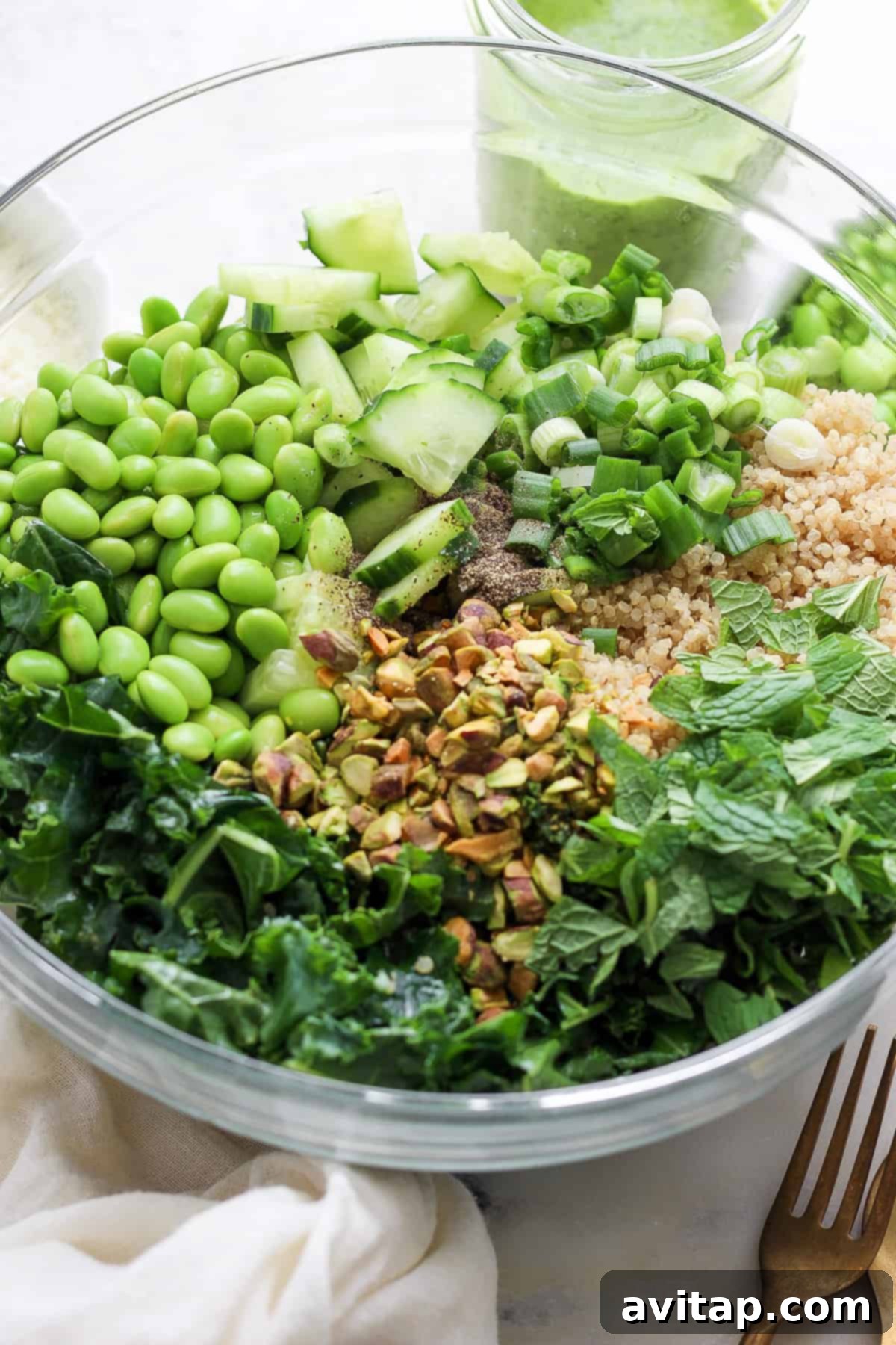 A large bowl brimming with a fresh, healthy Green Goddess Quinoa Salad, featuring a vibrant mix of fluffy cooked quinoa, tender massaged kale, crisp cucumber slices, bright green peas, crunchy chopped pistachios, and aromatic fresh herbs, all tossed together and ready for serving.