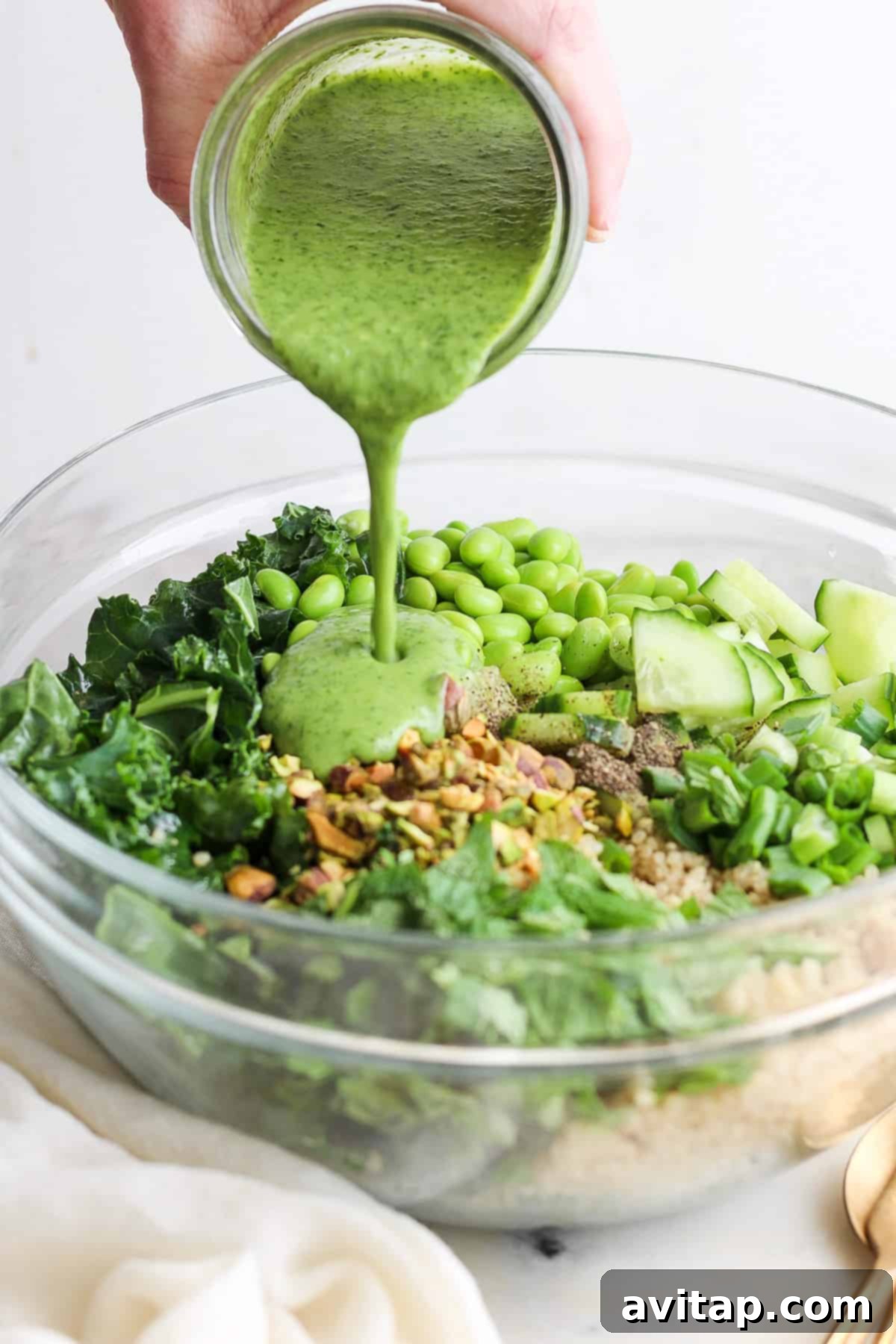 A generous serving of Green Goddess Quinoa Salad, featuring a base of tender kale and fluffy quinoa, with visible crisp cucumber slices and bright green peas, being richly drizzled with the creamy, vibrant green homemade dressing.