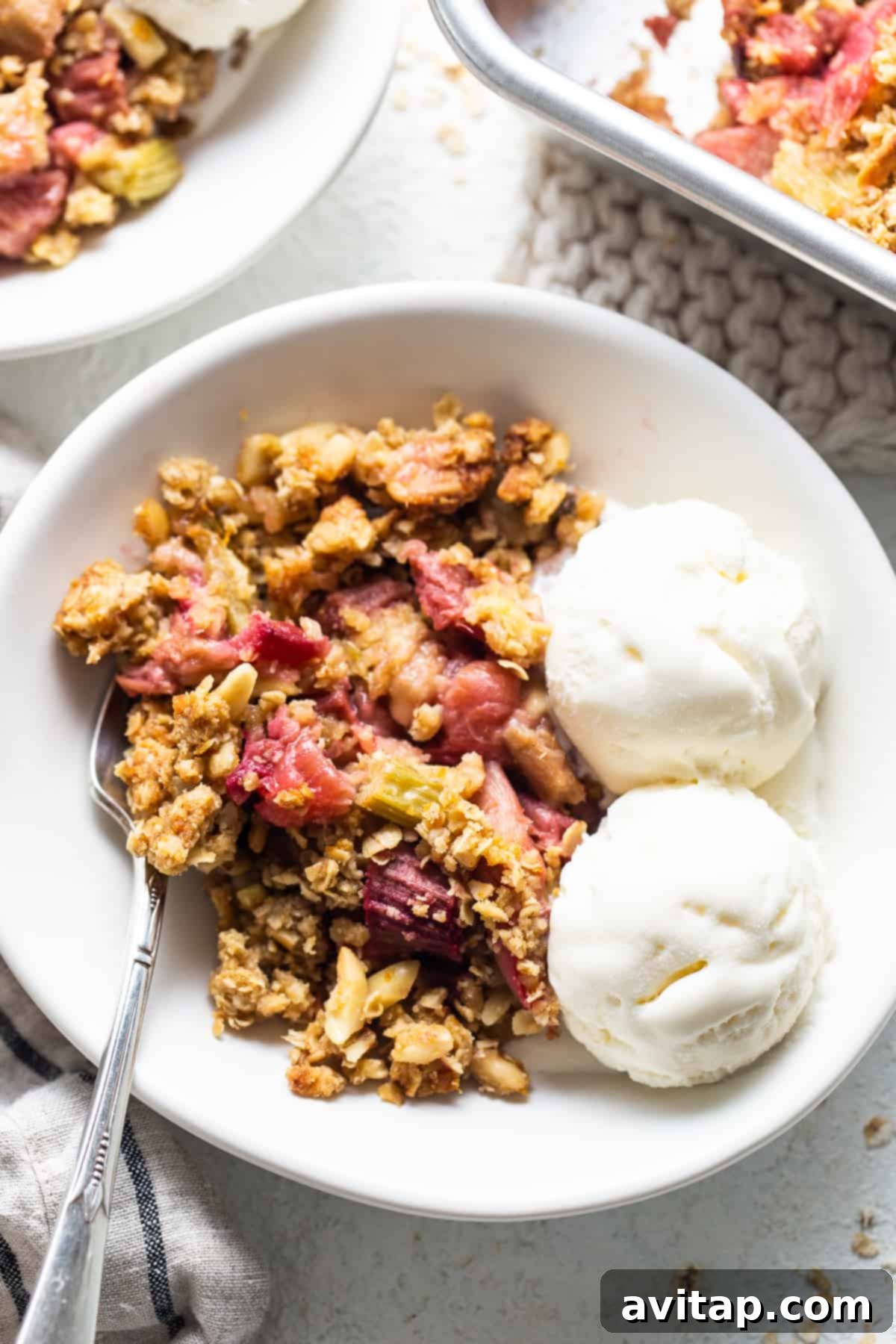A close-up shot of a bowl of warm rhubarb crisp, garnished with a generous scoop of vanilla ice cream and fresh berries, ready for serving.