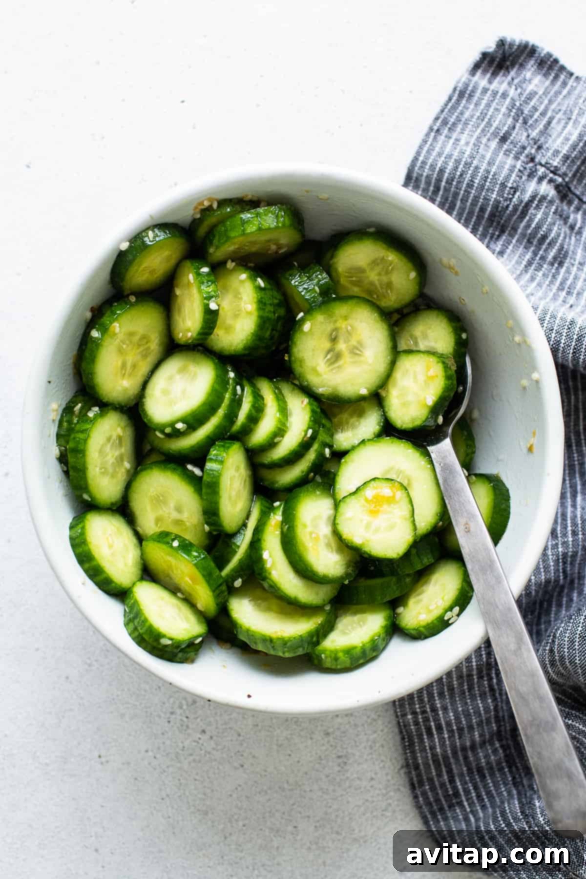 A crisp white bowl filled with vibrant green sliced mini cucumbers, tossed with a flavorful dressing, and a spoon for serving.