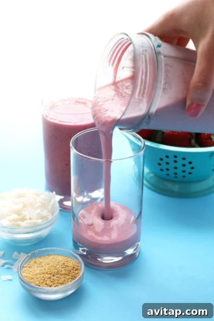 Strawberry Coconut Workout Refuel 3 A hand is shown expertly pouring a thick, creamy strawberry coconut protein smoothie from a blender into a tall glass, illustrating the perfect consistency of the drink.