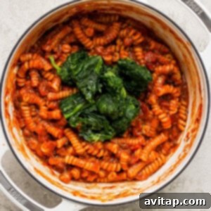 Pasta with tomato sauce and vibrant spinach in a white pot, nearing completion.