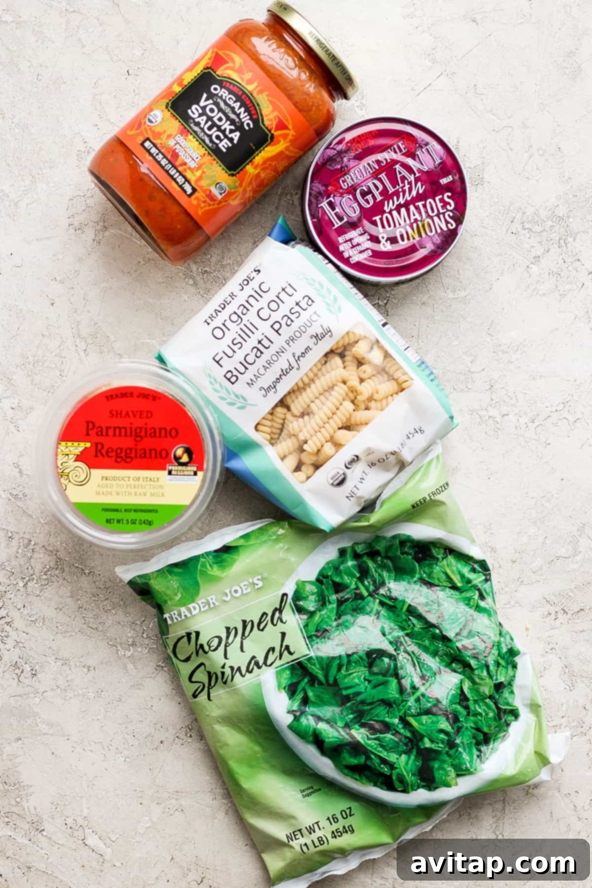 Easy One-Pot Pasta Alla Vodka 3 A curated selection of Trader Joe's grocery items for the pasta dish, including tomato sauce, canned eggplant, rigatoni pasta, chopped spinach, and parmesan cheese, neatly arranged on a bright surface.