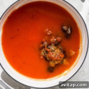 A pot containing a simmering tomato soup base with herbs and vegetables.