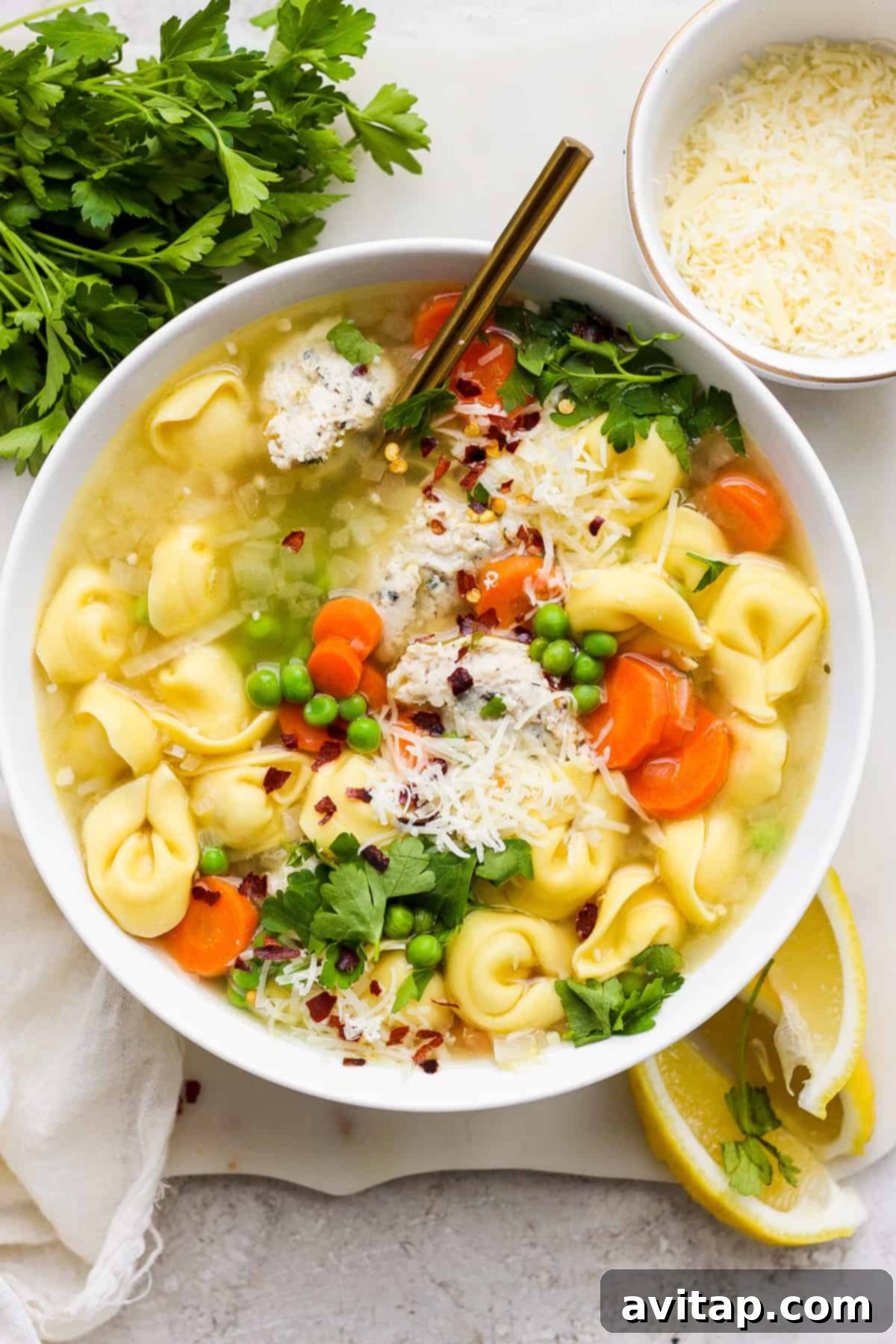 A bowl of chicken tortellini soup garnished with fresh parsley, shredded cheese, and a sprinkle of red pepper flakes, served with lemon wedges on the side.
