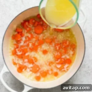 Pouring chicken broth into a pot with cooking onions and carrots.