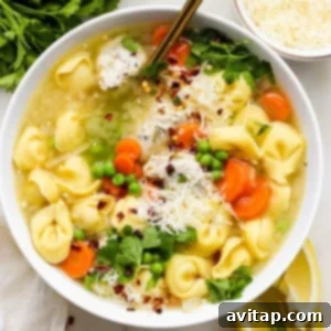 A bowl of chicken tortellini soup garnished with fresh parsley, shredded cheese, and a sprinkle of red pepper flakes, served with lemon wedges on the side.