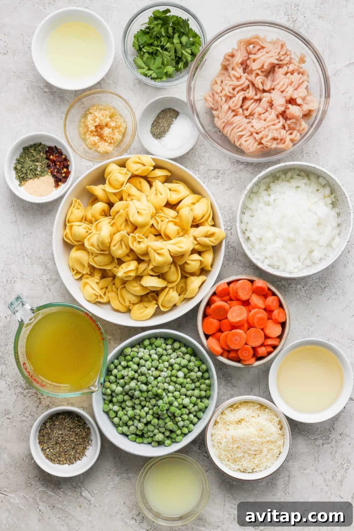 Assorted fresh ingredients for cooking laid out on a wooden surface, including pasta, a variety of colorful vegetables, ground meat, a selection of spices, and a container of broth.