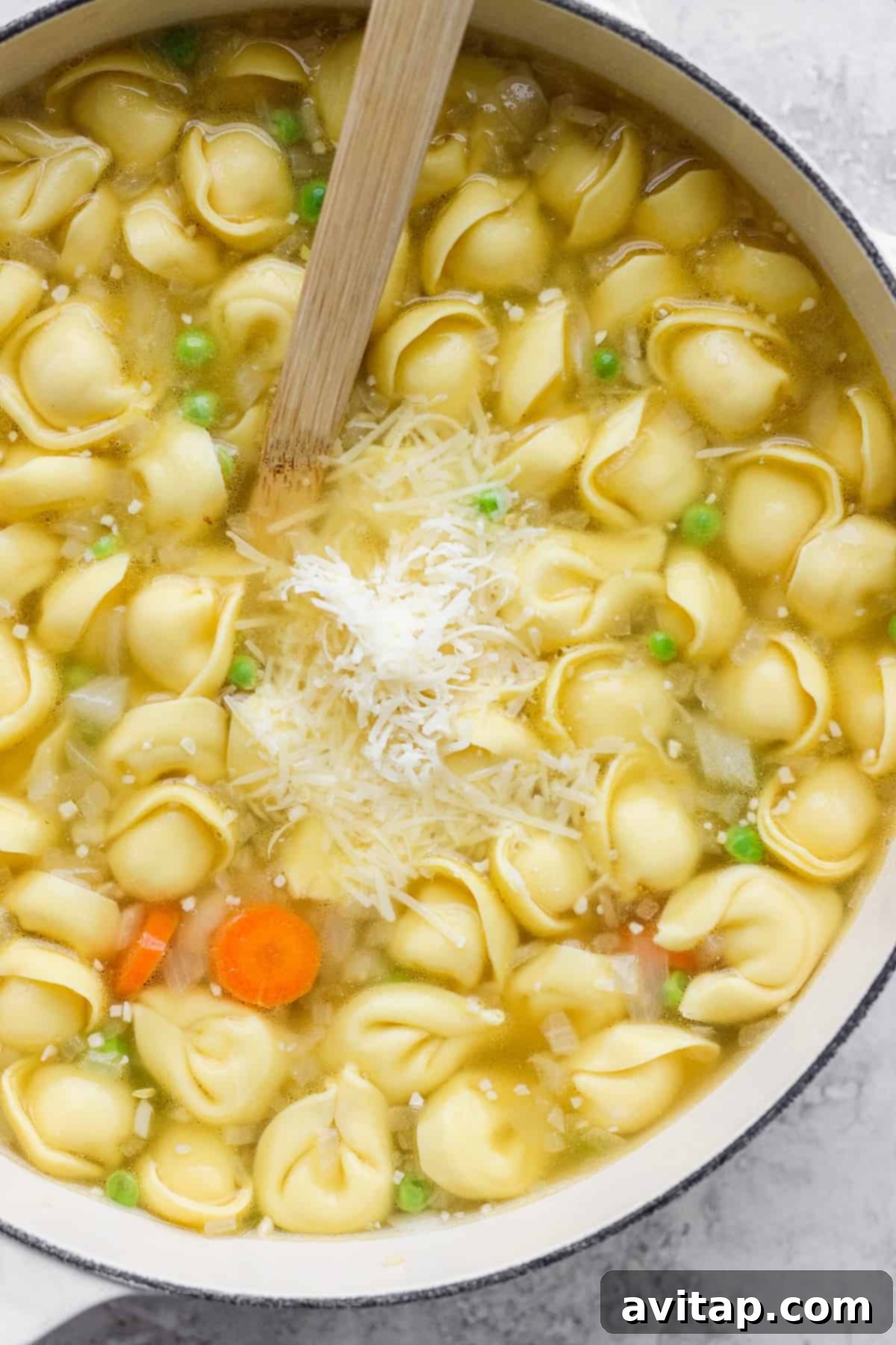 A large pot of chicken tortellini soup, featuring visible carrots and green peas, with a generous layer of grated cheese on top, ready to be served.