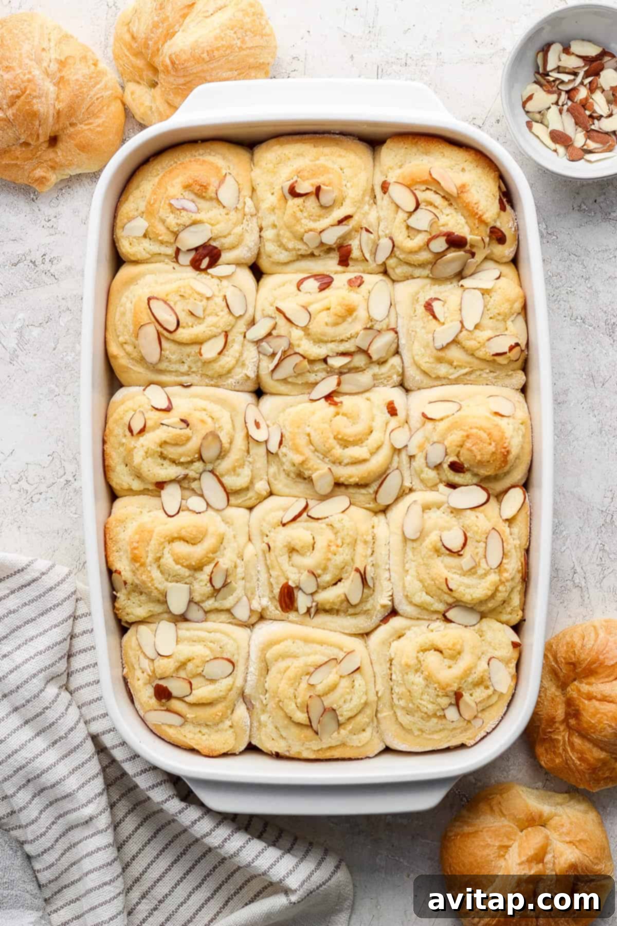 A freshly baked batch of Almond Croissant Cinnamon Rolls, generously topped with an almond cream filling and slivered almonds, in a white baking dish.