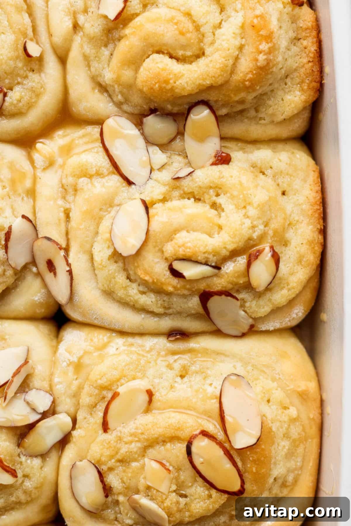 A freshly glazed Almond Croissant Cinnamon Roll, featuring a glistening white vanilla glaze over the golden-brown pastry and toasted almond slivers.