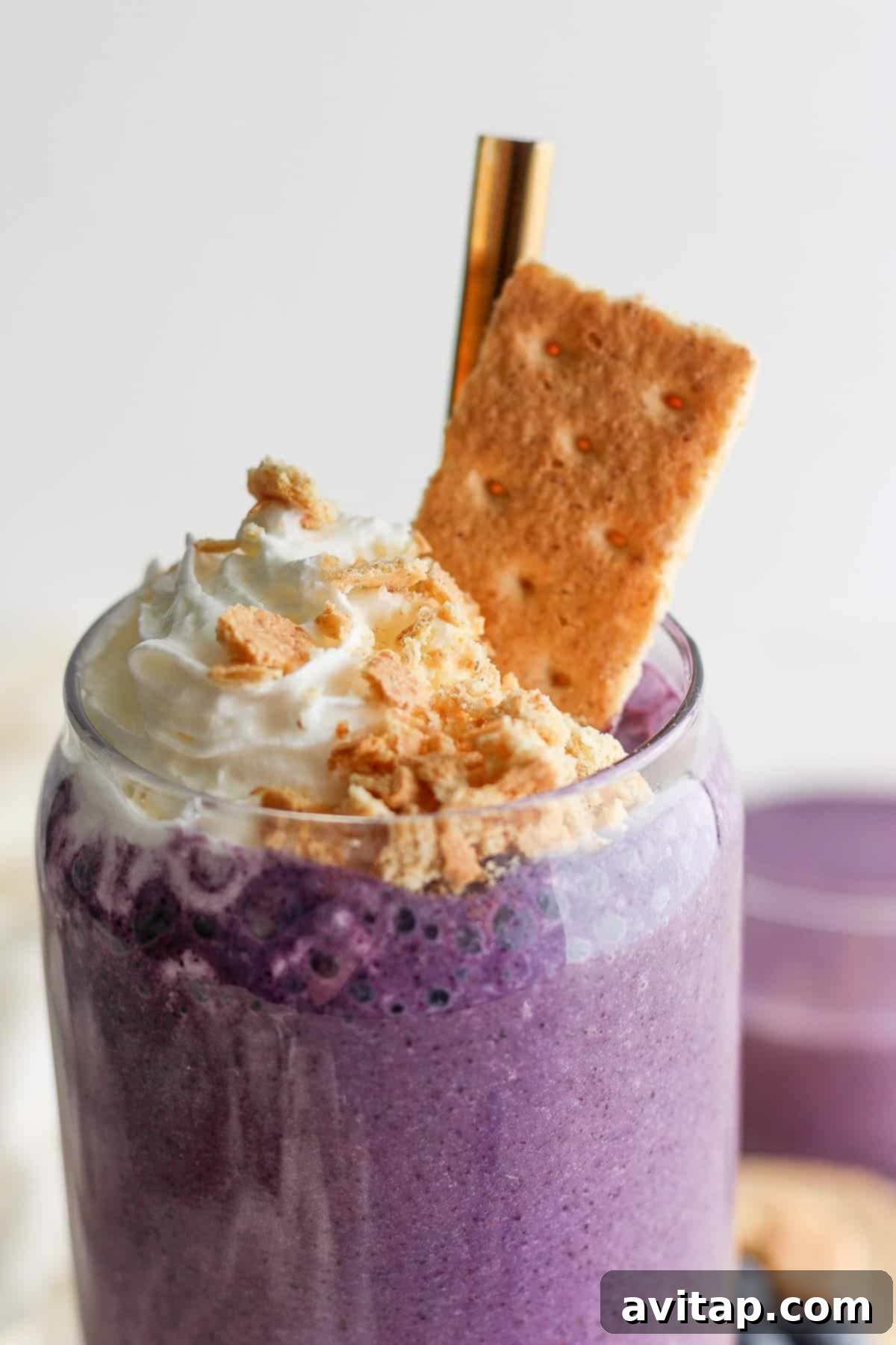 A beautifully presented vibrant purple blueberry cheesecake protein smoothie, topped with whipped cream and crushed biscuits, served in a clear glass with a elegant gold straw.