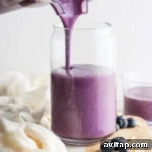 Pouring a fresh blueberry smoothie into a glass jar, surrounded by scattered blueberries on a wooden board, showcasing the finished product.